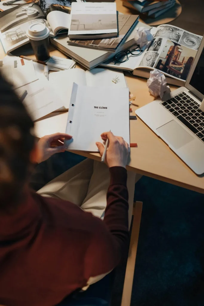 A person writing notes on a screenplay surrounded by books and a laptop on a work desk.